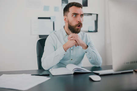young bearded amazed brunette man looking surprised while working on business project in his modern office, holds hands together , work routine conceptの写真素材