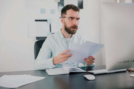 young bearded brunette man looking amazed while working on business project in his modern office, holding documents in his hand, work routine conceptの写真素材
