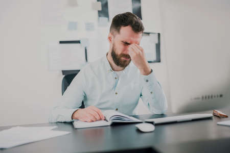 young bearded brunette man looking tired while working on business project in his modern office, holding his hand near face, work routine conceptの写真素材