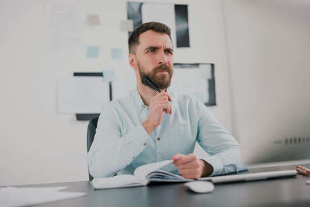 young bearded brunette man looking thoughtful while working on business project in his modern office, holding pen in his hand, work routine conceptの写真素材