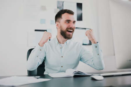 young bearded brunette man looking happy after successful business contract, sitting in his modern office with hands in winning position, success and well-being conceptの写真素材