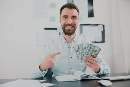 bearded man sitting in modern office holding cash in his hand like card deck looking excited while working on business project , money making motivation conceptの写真素材