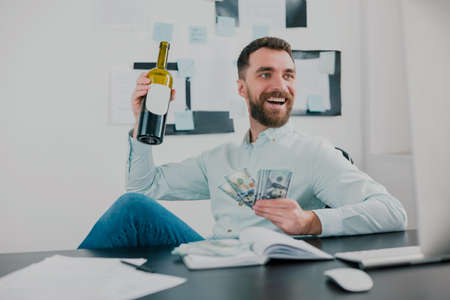bearded man drinking wine celebrating successful business deal in modern office holding cash in his hand like card deck looking excited, money making motivation conceptの写真素材