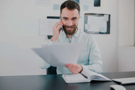 young bearded brunette man working on business project in his modern office, holding documents in his hand, talking on the phone, work routine conceptの写真素材