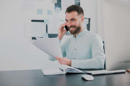 young bearded smiling man working on business project in his modern office, holding documents in his hand, having phone conversation, work routine conceptの写真素材