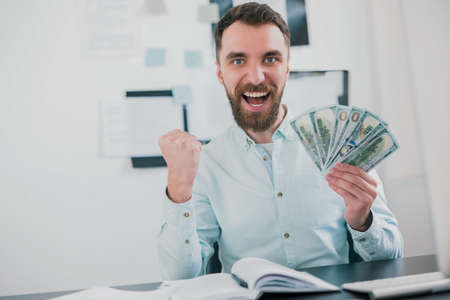 bearded man sitting in modern office holding cash in his hand like card deck looking holding his fist in winning position, money making motivation conceptの写真素材