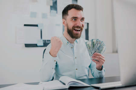 bearded man sitting in modern office holding cash in his hand like card deck looking excited while working in his office, money making motivation conceptの写真素材