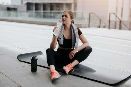 happy young beautiful brunette woman in sportswear warming up before street workout stretching on yoga mat, drinking water from the bottle, healthy fit lifestyleの写真素材