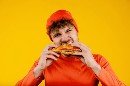 Young handsome hungry man bites sandwich in both hands on isolated yellow backgroundの写真素材