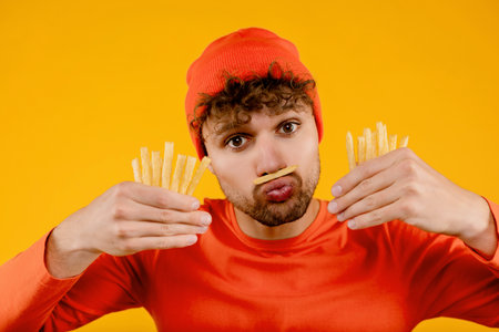Young funny man holds fried potatoes in both hands put one french fry on his mouth, isolated yellow backgroundの写真素材