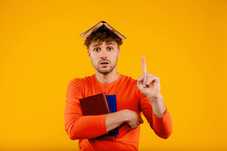 Young handsome surprised man with book on his head, points with finger up prepares for exams, on yellow backgroundの写真素材