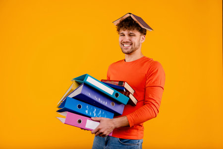 Young handsome happy man office worker with book on his head, holding document folders, isolated yellow backgroundの写真素材