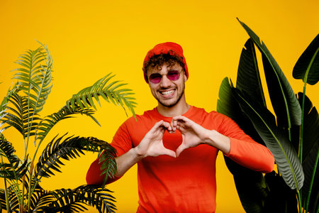 Portrait of young attractive man showing heart with his hands , love concept on yellow background with green plantsの写真素材
