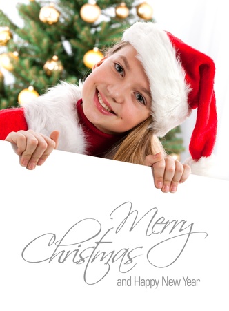 Pretty blond hair girl with santa hat and red comforter holding white board with space for text  Christmas tree in background の写真素材