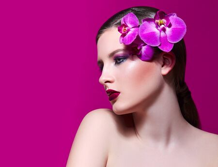 A beautiful girl is holding a lilac orchid in her hands in front of her face. Blue beautiful fresh natural flower in hands of the girl. Portrait of a girl with flowers in studio on a pink background.の写真素材