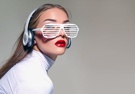 A beautiful girl in white glasses with big white headphones and loose hair, listening to music. A brown-haired woman with long straight hair with red lips in white robes. Studio, background.の写真素材