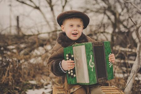 Boy in a hat and jacket with an accordion singing outdoors in winter. Happy young boy playing accordion.Little funny accordion player.の写真素材