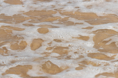 A sandy beach with a foamy, white wave crashing onto the shore. The foam is scattered across the sand, creating a dynamic and lively sceneの写真素材
