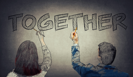 Young man and woman writing, the word together, with pencil on the grey wall. Business collaboration teamwork and growth. Strong partnership and foundation as a business conceptの写真素材