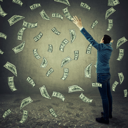 Rear View of a excited businessman with his hands raised up looking at falling money as a rain of dollars. Lottery winning, celebrating success. Symbol of wealth and good luck.の写真素材