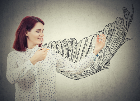 Young smiling businesswoman drawing a wing sketch behind her back. Create super power idea, self development success and freedom concept isolated on grey wall background.の写真素材