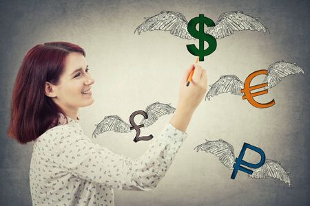 Young businesswoman holding a pencil in her hand, drawing winged money signs, as dollar, euro, pound and ruble are flying away isolated on gray wall background.の写真素材