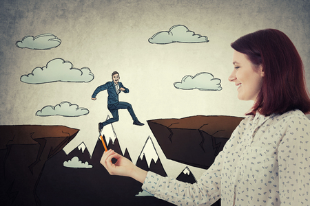 Smiling woman holding a pencil in her hand drawing a sketched businessman jumping over a chasm obstacle. Symbol of challenge and risk. Manipulation concept, business control as a marionette.の写真素材