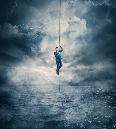 Businessman salvation, surviving the storm buiness concept as a scared man hanging on a chain above the ocean water, trying to climb up. Risk symbol, metaphor for conquering adversity and overcoming challenges.の写真素材