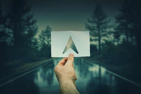 Close up of a man hand holding a white paper sheet with gps arrow icon inside over a foggy road between a forest background. Follow the correct way to success. Concept of the important choice in business.の写真素材