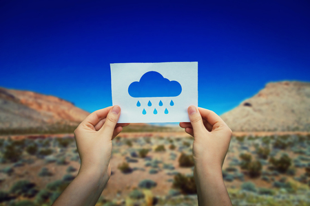 Close up of woman hands holding a white paper sheet with a rain cloud icon over a hot desert background. Dreaming of a rainy day in a sunny season.の写真素材