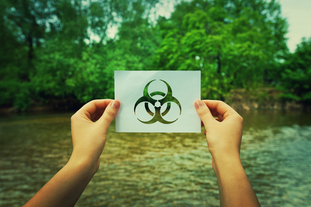 Woman hands holding a paper sheet with infection symbol in the middle of the nature near a river flowing and the forest. Biohazard sign, warning virus alert for hazardous epidemic bio materials.の写真素材