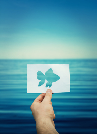 Man hand holding a white paper sheet with the golden fish shape on a sea shore background. Hope to catch the golden fish for fulfill his three wishes. Dream of success concept.の写真素材