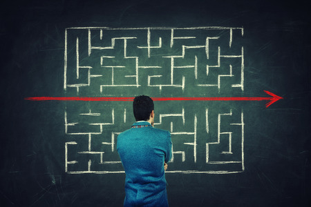 Rear view of a puzzled businessman in front of a blackboard finding a solution to escape from labyrinth. Breaking the rules, as a red line pierce the maze walls.の写真素材