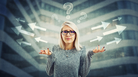 Confused young woman wearing glasses and frustrated hand gesture have to choose arrow direction,...