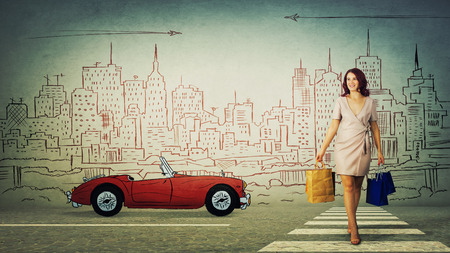 Full length portrait of beautiful young woman carrying shopping bags cross the road walking on zebra in a imaginary city with a retro race car sketch.の写真素材