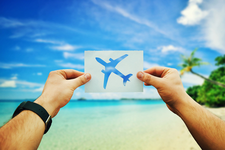 Close up of man hands holding a white paper sheet with airplane icon over tropical beach background with blue sea, clear sky and exotic palm trees.の写真素材