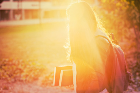 Silhouette of a young student woman carrying backpack and books, sun rays calm and peaceful orange effect.の写真素材