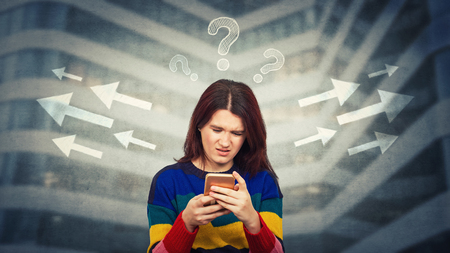 Woman looking at smartphone with puzzled face confused and frustrated has to choose arrow direction, left or right side, interrogation marks above head. Difficult decision concept, correct choice.の写真素材