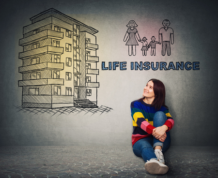 Young woman sitting on the floor smiling looking at a flat house and happy family sketch on the wall. Life insurance, family and home protection concept.の写真素材