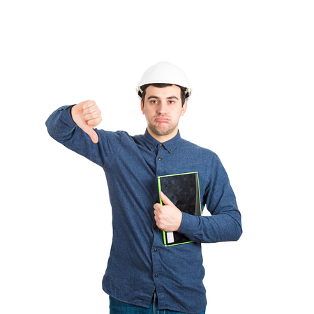 Unhappy and tired young man engineer wearing protective helmet holding tablet computer showing thumb...
