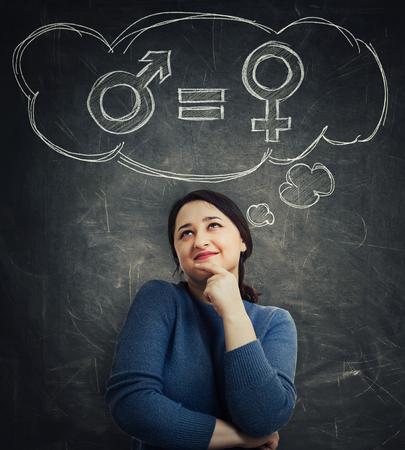 Casual young woman holding hand under chin looking up dreaming as a thought cloud showing gender equality concept with male and female symbol over blackboard background. Sex sign social issue metaphorの写真素材