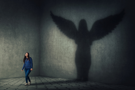 Proud and confident young woman walking and casting a superhero shadow as a guardian angel with wings on a dark room wall. Inner power, ambition and leadership concept.の写真素材