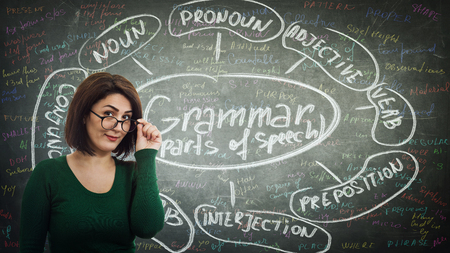 Young woman teacher fixing glasses looking to camera in front of blackboard written with chalk english grammar parts of speechの写真素材