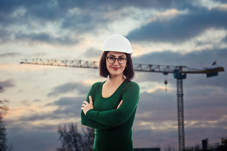 Beautiful confident woman engineer positive smiling wearing glasses and protective helmet, holding arms crossed, over constructions site at the back with a tall tower crane and sunset sky.の写真素材