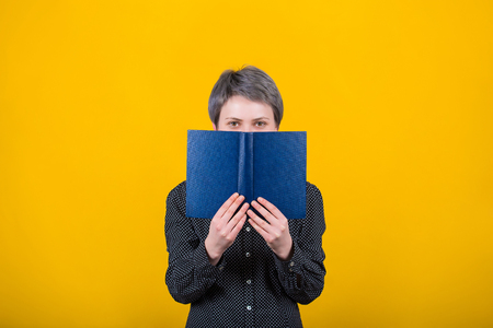 Happy woman book reader hides behind a blue textbook, has joyful expression and positive sight,...