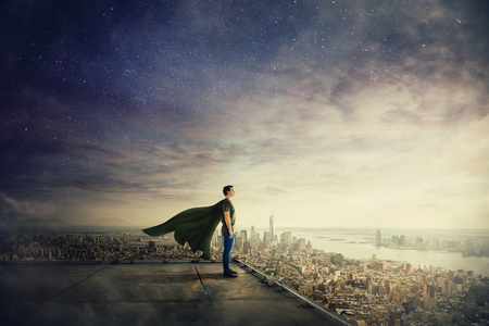Surreal view of confident superhero with cape stands on the rooftop looking over city horizon. Ambition and business success concept. Leadership hero power, motivation and inner strength symbol.の写真素材