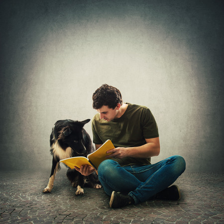 Curious purebred border collie dog and his master reading together an interesting book. Owner seated on floor training his cute puppy isolated over grey wall. Human and animal friendship concept.の写真素材
