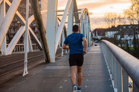 Young athletic man practicing sport outdoors, running fast along bridge over the sunset sky...