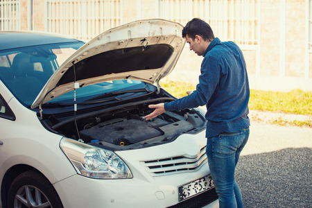 Perplexed young male driver, shrugging shoulders, looking confused under the popped hood of his car. Broken transportation, vehicles problems and breakdowns concept. Guy needs help with towing.の写真素材