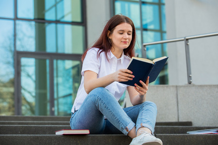 University student outdoors on Campus sitting on stairs holding books. A confident student holding books over university building background learning for an exam.の写真素材